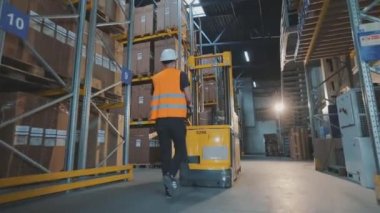 Bir işçi depodan kargo taşıyor. Bir işçi elektrikli forkliftle depoya giriyor..