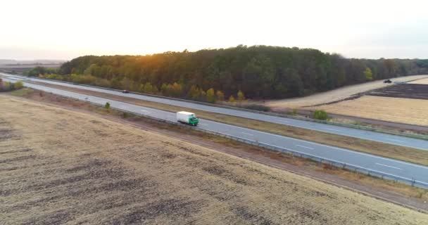 Lastbilar kör längs motorvägen bland fälten vid solnedgången. Lastbilar färdas på en rak väg vid solnedgången ovanifrån — Stockvideo