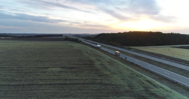 Lastbilar färdas på en rak väg vid solnedgången ovanifrån. Lastbilar kör längs motorvägen bland fälten vid solnedgången. — Stockvideo