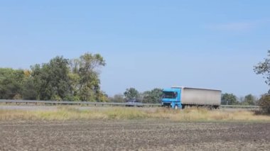 Güneşli bir havada beyaz bir araba otoban boyunca sürer. Otoyolda kamyon var.