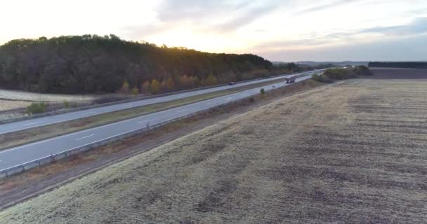 Ciężarówki jechać wzdłuż autostrady wśród pól o zachodzie słońca. Ciężarówki podróżują po prostej drodze o zachodzie słońca widok z góry — Wideo stockowe