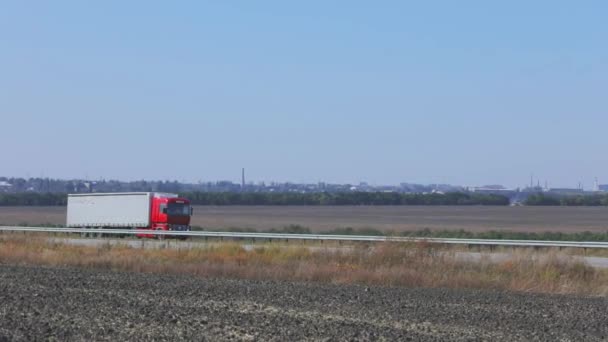 Ciężarówka z czerwoną kabiną jedzie wzdłuż autostrady. Ciężarówka jedzie wzdłuż drogi. Dostawa towarów ciężarówką — Wideo stockowe