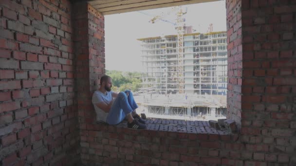 Un jeune homme s'assoit à la fenêtre sur le fond d'un chantier et regarde le téléphone. Un jeune homme chez un ouvrier de la construction regarde le téléphone. Il est assis sur la fenêtre de la maison. — Video