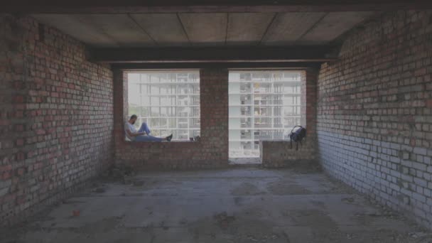 En ung man sitter vid fönstret mot bakgrunden av en byggarbetsplats och tittar på telefonen. En ung man på en byggarbetare tittar på telefonen. Killen sitter på fönstret i huset — Stockvideo