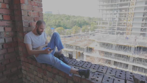 那家伙坐在房子的窗户上。一个年轻人坐在建筑工地的窗前，看着电话。一个在建筑工人面前的年轻人看着电话. — 图库视频影像