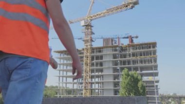 İnşaat alanındaki iki mühendis çizime bakıyor. Mühendisler çok katlı bir binanın önünde. Evin arka planında iki inşaatçı var.