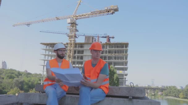 Μηχανικοί μπροστά από ένα πολυώροφο κτίριο. Δύο μηχανικοί σε ένα εργοτάξιο κοιτάζουν το σχέδιο. Δύο χτίστες στο βάθος του σπιτιού. — Αρχείο Βίντεο