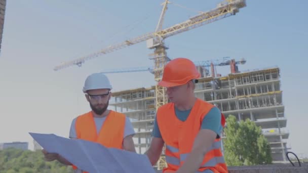 Twee ingenieurs op een bouwplaats kijken naar de tekening. Ingenieurs voor een gebouw met meerdere verdiepingen. Twee bouwers op de achtergrond van het huis — Stockvideo