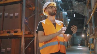 Modern bir depoda bir adam. Depo müdürü depoyu kontrol ediyor. Depoda kask takan bir işçi.