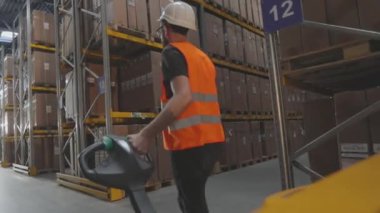 Bir işçi elektrikli forkliftle depoya doğru yürür. Bir işçi depodan kargo taşıyor.