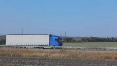 Güneşli bir havada beyaz bir araba otoban boyunca sürer. Otoyolda kamyon var.