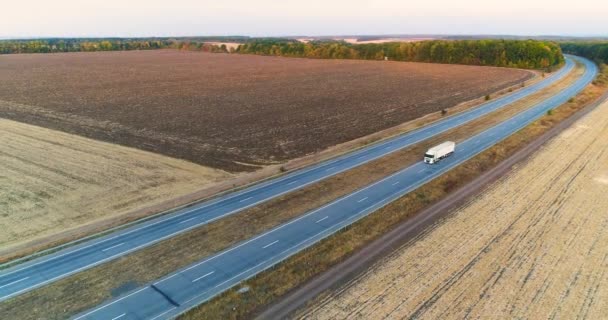 Camion guidano lungo l'autostrada tra i campi al tramonto. Camion viaggiano su una strada diritta al tramonto vista dall'alto — Video Stock