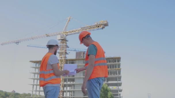 Ingenieure vor einem mehrstöckigen Gebäude. Zwei Ingenieure einer Baustelle betrachten die Zeichnung. Zwei Bauarbeiter im Hintergrund des Hauses — Stockvideo