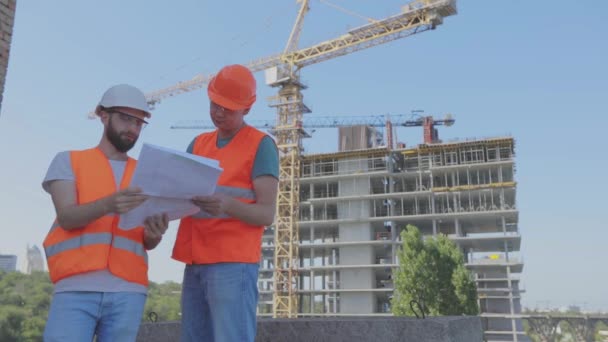 Dos ingenieros en un sitio de construcción están mirando el dibujo. Ingenieros frente a un edificio de varios pisos. Dos constructores en el fondo de la casa — Vídeo de stock