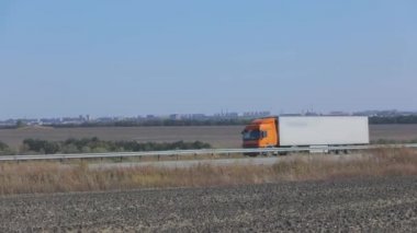 Güneşli bir havada beyaz bir araba otoban boyunca sürer. Otoyolda kamyon var.