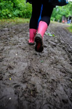 Bir müzik festivalinde çamurlu, asfaltsız bir yolda pembe lastik çizmeler giyen genç bir bayanla sığ alan derinliği (seçici odak).