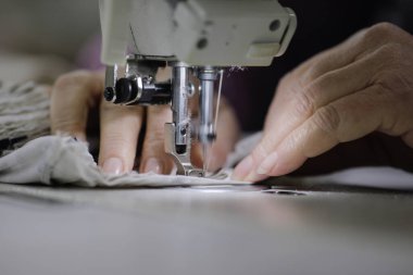 Bir tekstil fabrikasında elektrikli dikiş makinesinde çalışan yaşlı bir kadının elleriyle sığ alan derinliği (seçici odak) görüntüsü.