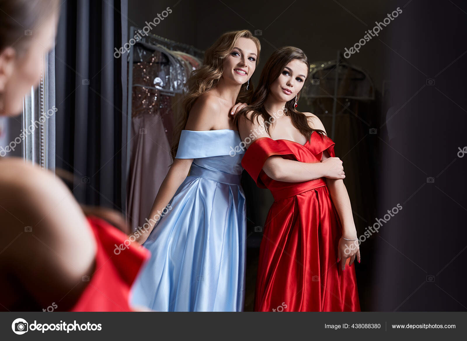 Two young beautiful girls wearing off-the-shoulder full-length sky blue and  crimson red satin slit prom ball gowns. Models looking in mirror. Fitting  room in dress hire service. Stock Photo by ©oleksandrberezko 444091090