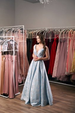 Genç, güzel, esmer bir kız. Uzun boylu, gökyüzü mavisi, saten yırtık balo elbisesi giyiyor. Üzerinde de işlemeli gül deseni var. Elbise kiralama servisi ve arka planda birçok elbise..