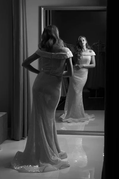 若い美しいブロンドの女の子は、フル長のシフォンの人魚のドレスやスパンコールで飾られたプロムボールのガウンを身に着けています。フィッティングルームで鏡の前のモデル。黒と白の古典的なスタイル. — ストック写真
