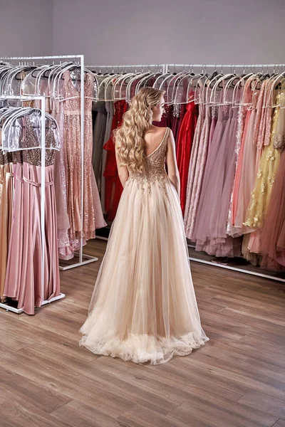 Young beautiful blonde girl wearing a full-length beige yellow champaign chiffon slit prom ball gown decorated with golden sparkles and sequins. Fashion model in dress hire service with many dresses.