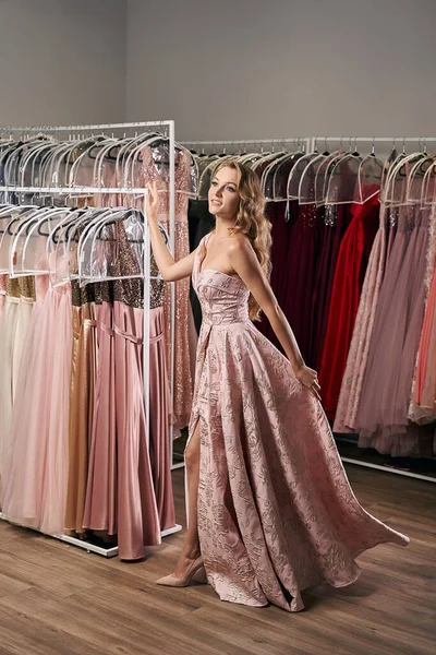 Joven hermosa chica rubia con una longitud completa cubierta de un hombro vestido de baile de raso rosa pálido raja decorado con patrón bordado. Servicio de alquiler de vestidos con muchos vestidos en el fondo. — Foto de Stock