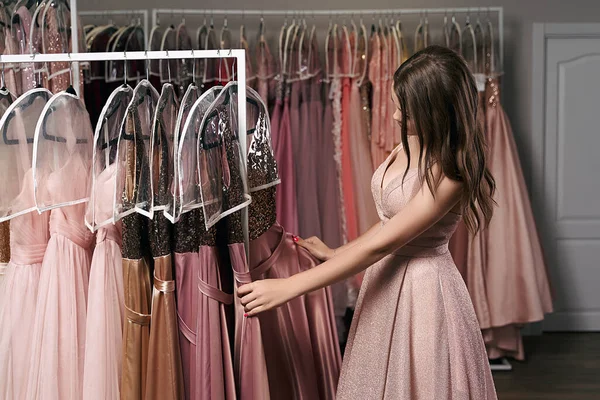 Portrait of young beautiful girl wearing a full-length pale pink glitter chiffon draped prom ball gown. Model selecting an outfit for occasion in dress hire service with many options on background. — Stock Photo, Image