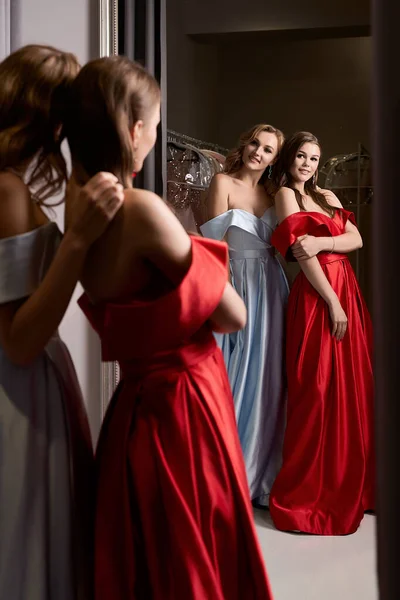 Duas jovens meninas bonitas vestindo off-the-ombro completo céu azul e vermelho carmesim fenda de cetim vestido de baile de formatura. Modelos a olhar ao espelho. Quarto de montagem em serviço de aluguer de vestido. — Fotografia de Stock