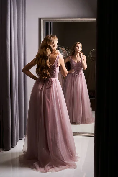 Young beautiful blonde girl wearing a full-length pink violet or purple chiffon prom ball gown decorated with sparkles and sequins. Model in front of mirror in a fitting room at dress hire service. — Stock Photo, Image