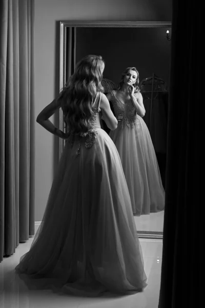 Young beautiful blonde girl wearing a full-length pink violet or purple chiffon prom ball gown decorated with sparkles and sequins. Model in front of mirror in a fitting room. Black and white. — Stock Photo, Image