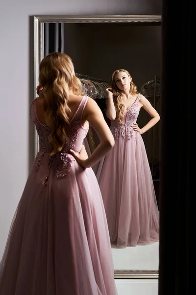 Menina loira bonita nova vestindo um vestido de baile de formatura de chiffon violeta ou roxo de comprimento total decorado com brilhos e lantejoulas. Modelo na frente do espelho em um quarto de montagem no serviço de aluguer de vestido. — Fotografia de Stock