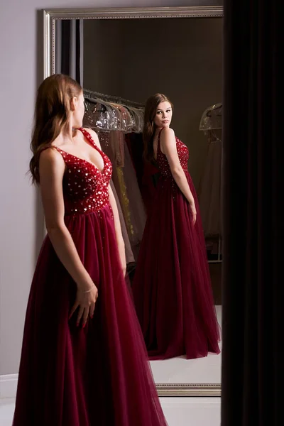 Menina morena bonita nova vestindo um full-length escuro vermelho chiffon vestido de baile de formatura decorado com brilhos e lantejoulas. Modelo na frente do espelho em um quarto de montagem no serviço de aluguer de vestido. — Fotografia de Stock