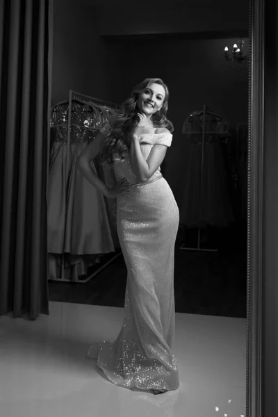 Young beautiful blonde girl wearing a full-length chiffon mermaid dress or prom ball gown decorated with sequins. Model in front of mirror in a fitting room. Black and white classic style. — Stock Photo, Image