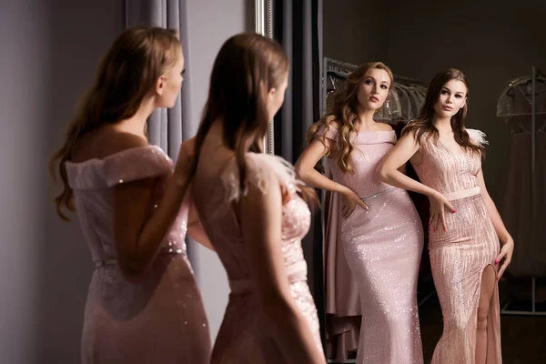 Dos chicas hermosas jóvenes que usan vestidos de sirena de gasa rosa o púrpura o vestidos de fiesta decorados con brillantes y lentejuelas. Modelos delante del espejo en un probador. — Foto de Stock
