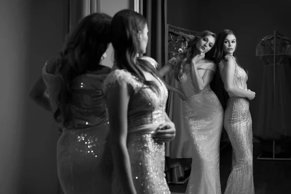 Two young beautiful girls wearing full-length pink violet or purple chiffon mermaid dresses or prom ball gowns decorated with sparkles and sequins. Models in front of mirror in a fitting room. — Stock Photo, Image