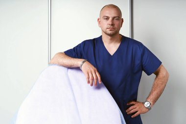 Profesyonel üniformalı, masaj masasının yanında duran deneyimli bir erkek masaj terapisti ya da masör..