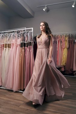 Mezuniyet balosu elbisesiyle süslenmiş açık pembe bir şifon giyen güzel bir kızın portresi. Model elbise kiralama hizmetinde arka planda birçok seçeneği olan bir kıyafet seçiyor.