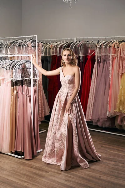 Young beautiful blonde girl wearing a full-length draped one shoulder pale pink satin slit prom ball gown decorated with embroindered pattern. Dress rental service with many dresses in on background.
