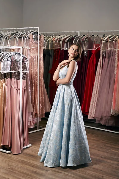 Young beautiful brunette girl wearing a full-length draped sky blue satin slit prom ball gown decorated with embroindered roses pattern. Dress hire service with many dresses on background.