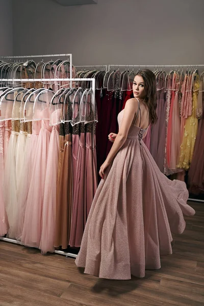 Retrato de menina bonita jovem vestindo um chiffon glitter rosa pálido de comprimento total drapeado vestido de baile de formatura. Modelo selecionando uma roupa para ocasião no serviço de aluguer de vestido com muitas opções em segundo plano. — Fotografia de Stock