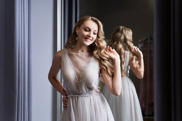 Young beautiful blonde girl wearing a full-length silver white chiffon prom ball gown decorated with sparkles and sequins. Model in front of mirror in a fitting room at dress hire service. — Stock Photo, Image