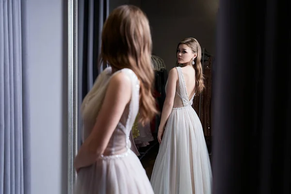 Joven chica morena hermosa que lleva un vestido de fiesta de gasa blanco plateado de cuerpo entero decorado con brillantes y lentejuelas. Modelo delante del espejo en un probador en el servicio de alquiler de vestidos. — Foto de Stock