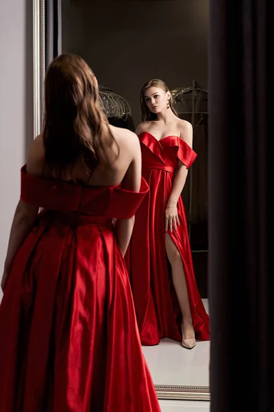 Young beautiful girl wearing an off-the-shoulder full-length crimson red satin slit prom ball gown. Model looking in mirror. Fitting room in dress hire service. — Stock Photo, Image