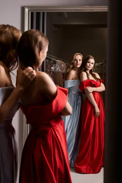 Две юные красавицы, одетые с плеча в бальные платья из красного атласа и голубого неба. Модели смотрят в зеркало. Тренажерный зал в аренду одежды. — стоковое фото