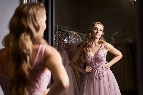Young beautiful blonde girl wearing a full-length pink violet or purple chiffon prom ball gown decorated with sparkles and sequins. Model in front of mirror in a fitting room at dress hire service. — Stock Photo, Image