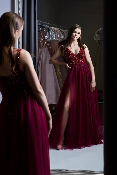 Młoda, piękna brunetka, ubrana w ciemnoczerwoną koszulę balową z szyfonem, ozdobioną błyszczącymi i cekinami. Model przed lustrem w przymierzalni w wypożyczalni sukni. — Zdjęcie stockowe