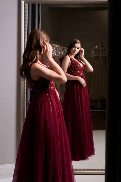 Menina morena bonita nova vestindo um full-length escuro vermelho chiffon vestido de baile de formatura decorado com brilhos e lantejoulas. Modelo na frente do espelho em um quarto de montagem no serviço de aluguer de vestido. — Fotografia de Stock