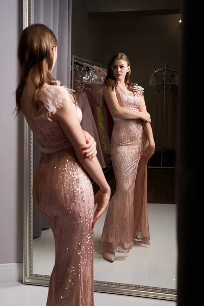 Junge schöne brünette Mädchen tragen ein in voller Länge blassrosa Passform Chiffon Ball Schlitz Kleid mit Funkeln und Pailletten verziert. Model vor Spiegel in Umkleidekabine. — Stockfoto