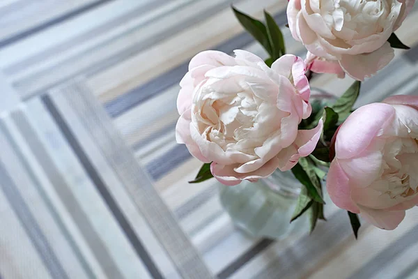 A bouquet of peonies in a transparent glass vase. Pink and white peonies on the table covered with pattern serving mats