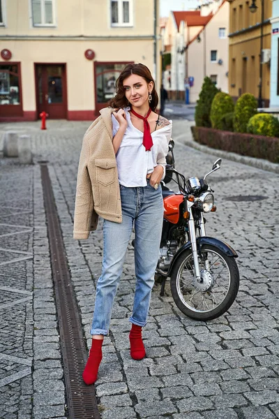 Mladá krásná žena v červené pletené tkaniny blok podpatek ponožky boty, modré džíny kalhoty a teddy bunda pózování v blízkosti motocyklu na ulici — Stock fotografie